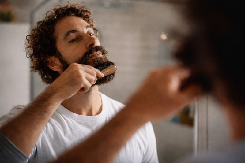 Man brushing his beard with a wooden brush while looking in the bathroom mirror.